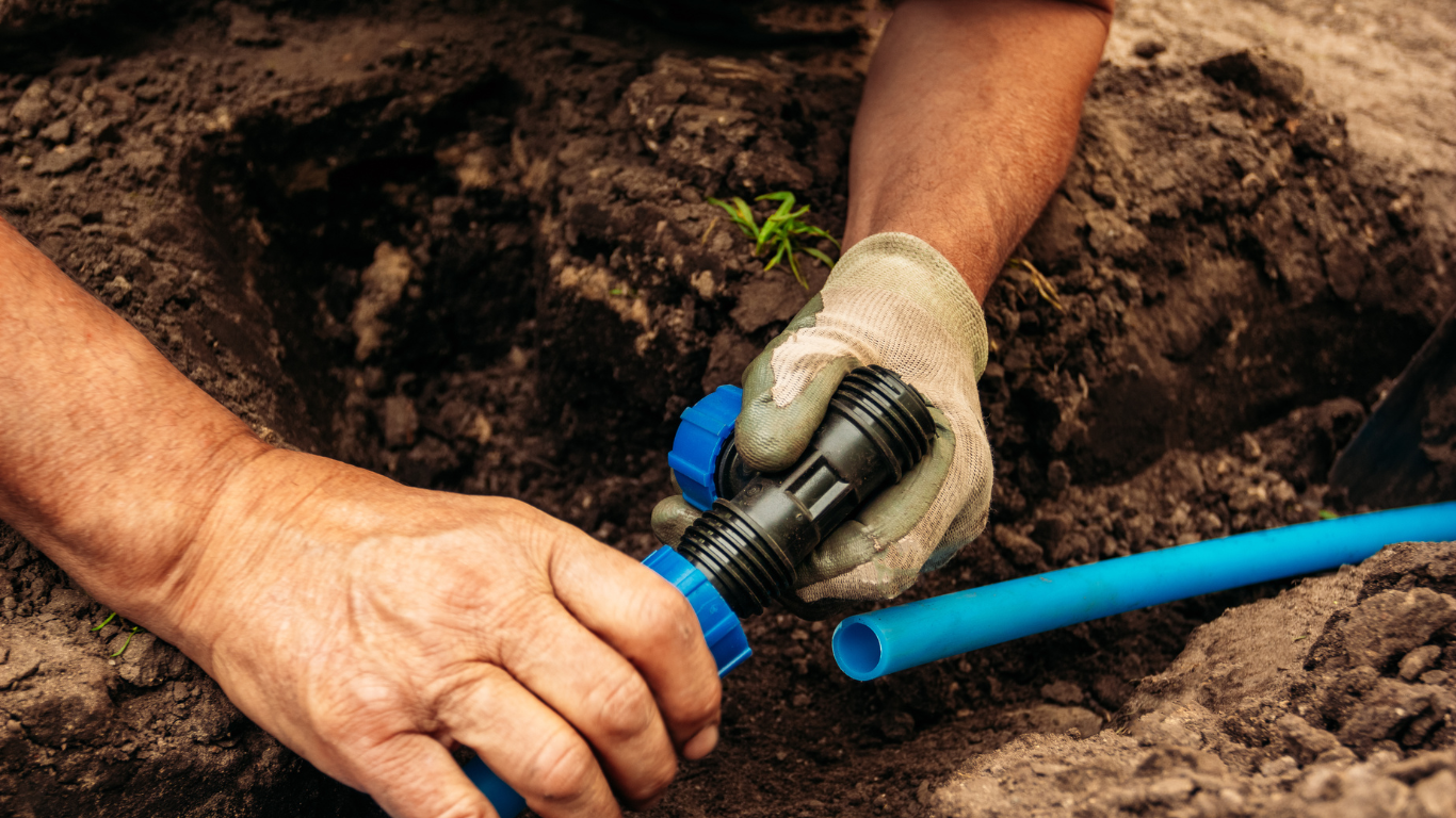Close-up of hands performing irrigation maintenance by connecting blue water pipes with a plastic fitting in a garden setting.